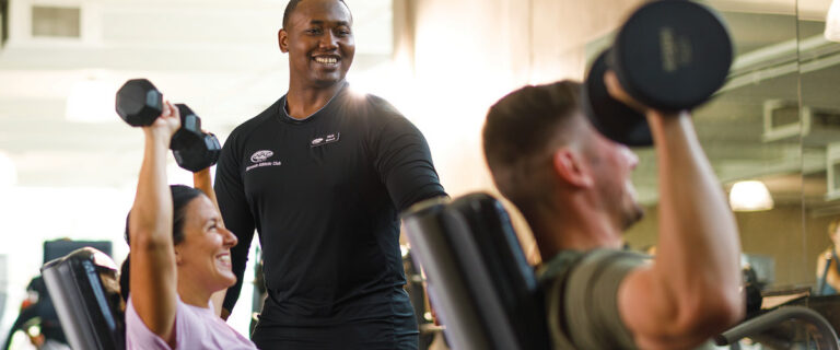 Smiling personal trainer works with a client couple lifting free weights